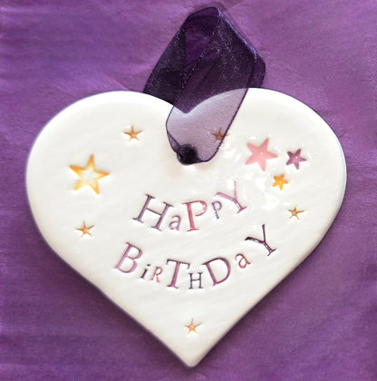 White ceramic heart with the words "Happy Birthday" and some small star shaped illustrations dotted around. There is a purple ribbon at the top of the heart for hanging.