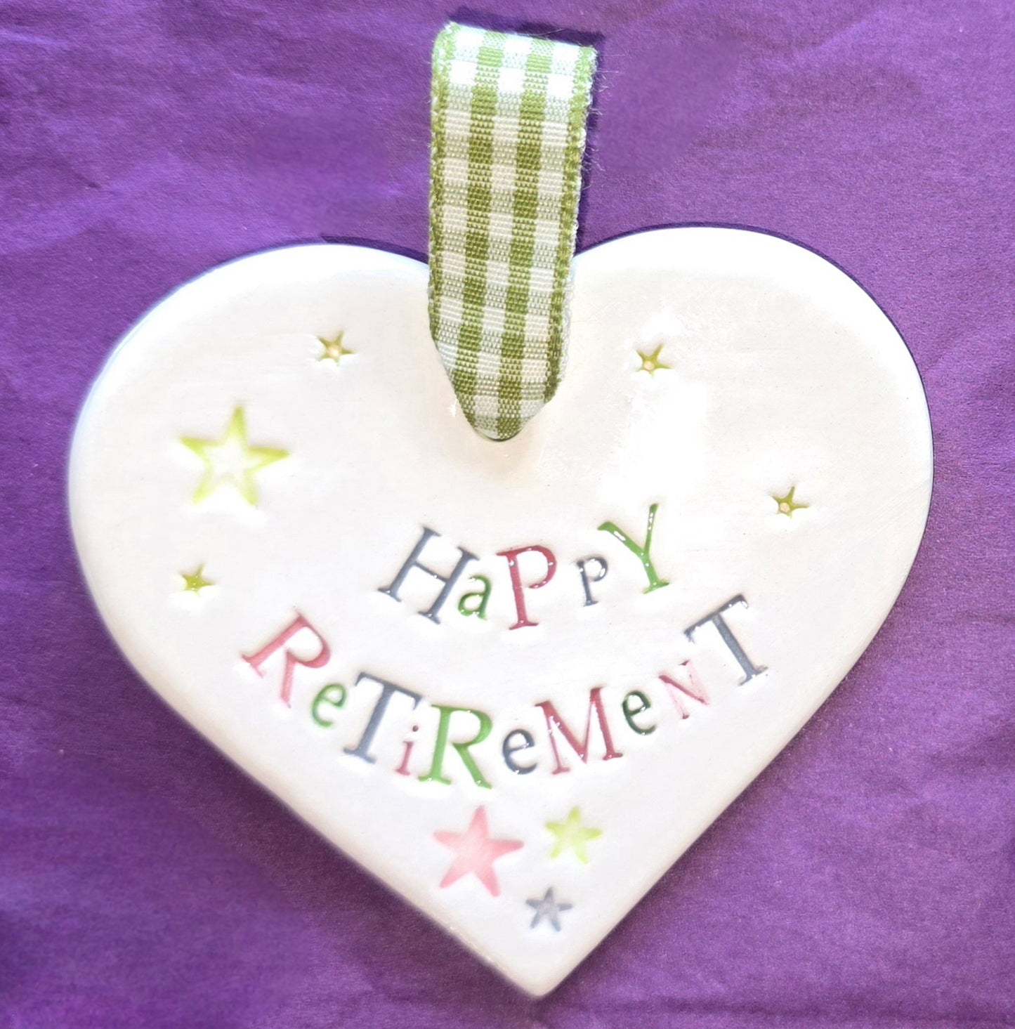 White ceramic heart with the words "Happy Retirement" and some small star shaped illustrations dotted around. There is a green checked ribbon at the top of the heart for hanging.