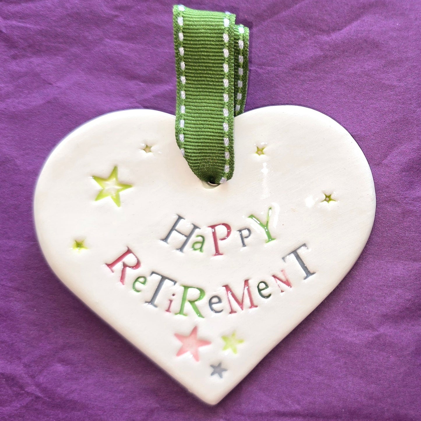 White ceramic heart with the words "Happy Retirement" and some small star shaped illustrations dotted around. There is a green ribbon at the top of the heart for hanging.