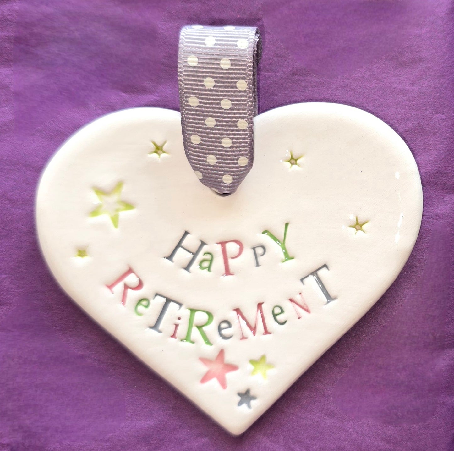 White ceramic heart with the words "Happy Retirement" and some small star shaped illustrations dotted around. There is a grey polka dot ribbon at the top of the heart for hanging.