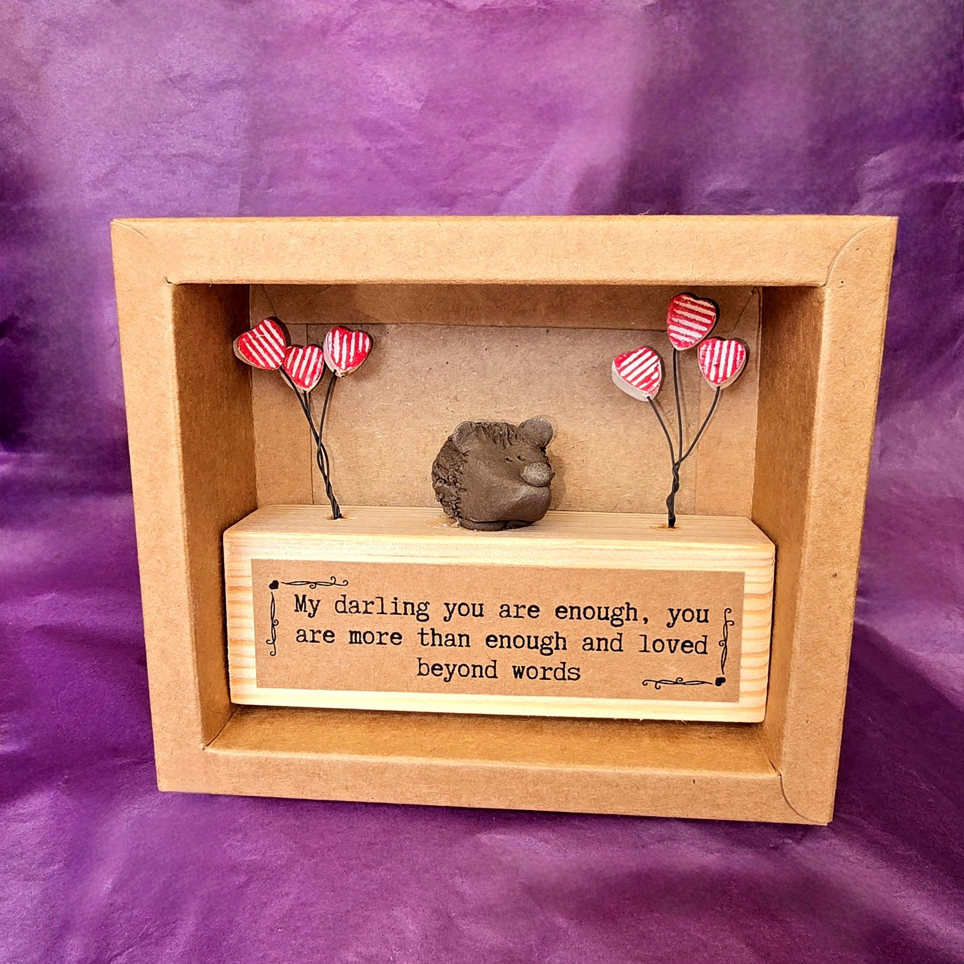 Open brown cardboard box containing the mini sculpture. Mini sculpture is made from a wooden block with a ceramic hedgehog sitting on top. To each side of the hedgehog there are three red and white striped hearts attached to wire. On the front of the wooden block there is a brown label with the text "My darling you are enough, you are more than enough and loved beyond words."