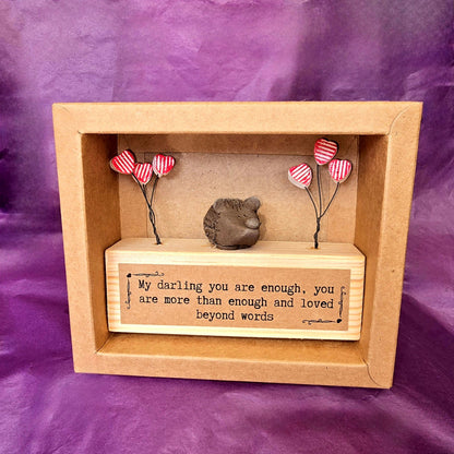 Open brown cardboard box containing the mini sculpture. Mini sculpture is made from a wooden block with a ceramic hedgehog sitting on top. To each side of the hedgehog there are three red and white striped hearts attached to wire. On the front of the wooden block there is a brown label with the text "My darling you are enough, you are more than enough and loved beyond words."