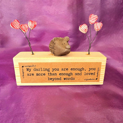 Mini sculpture made from a wooden block with a ceramic hedgehog sitting on top. To each side of the hedgehog there are three red and white striped hearts attached to wire. On the front of the wooden block there is a brown label with the text "My darling you are enough, you are more than enough and loved beyond words."