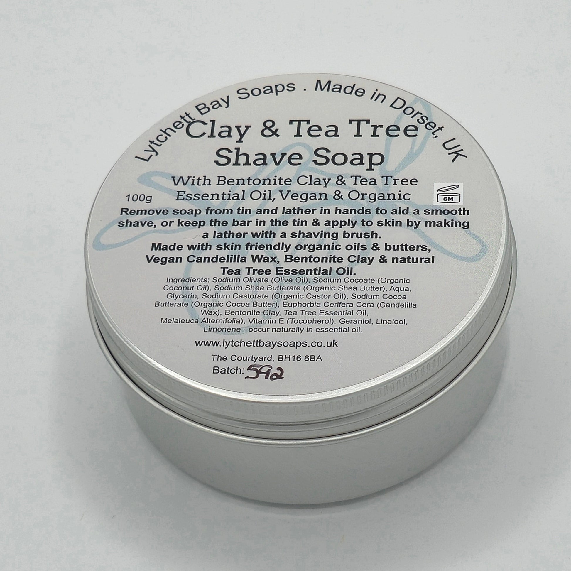 Round silver tin with light coloured label with the text "Lytchett Bay Soaps, made in Dorset, UK", "Clay & Tea Tree Shave Soap with Bentonite Clay & Tea Tree Essential Oil, Vegan & Organic". 
"Remove soap from tin and lather in hands to aid a smooth shave, or keep the bar in the tin & apply to skin by making a lather with a shaving brush. Made with skin friendly organic oils & butters, Vegan Candelilla Wax, Bentonite Clay & natural Tea Tree Essential Oil".