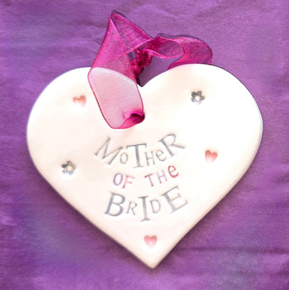 White ceramic heart with the words "Mother of the Bride" and some small heart and flower shaped illustrations dotted around. There is a burgundy ribbon at the top of the heart for hanging.