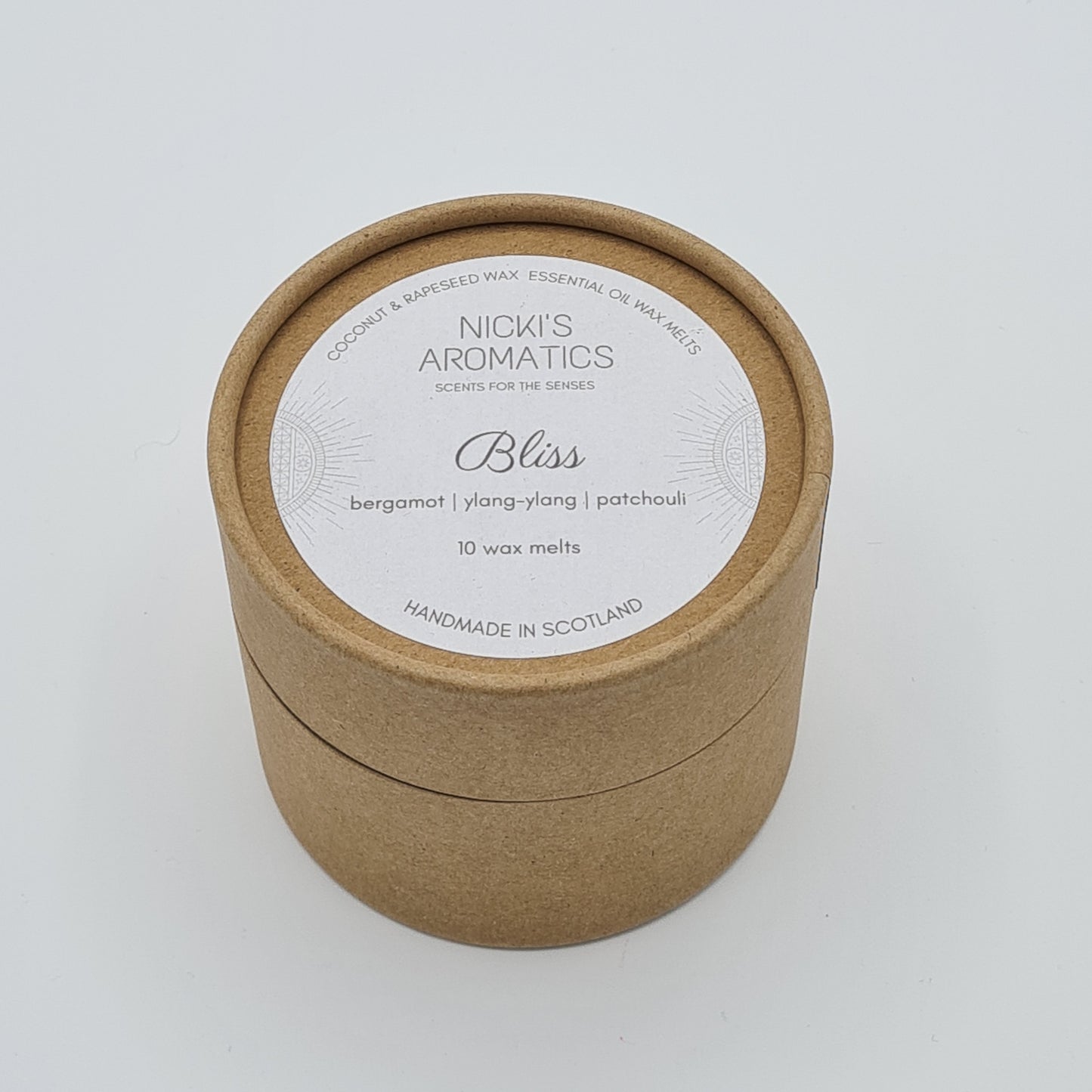 Round kraft cardboard box with a white label on the top with the text "Coconut & Rapeseed Wax Essential Oil Wax Melts" along the top. Below this is the logo for "Nicki's Aromatics" and the strapline "Scents for the Senses". In the middle of the label is the name "Bliss" with "Bergamot", "Ylang-Ylang" "Patchouli" and "10 wax melts" typed below. At the very bottom of the label is the wording "Handmade in Scotland".