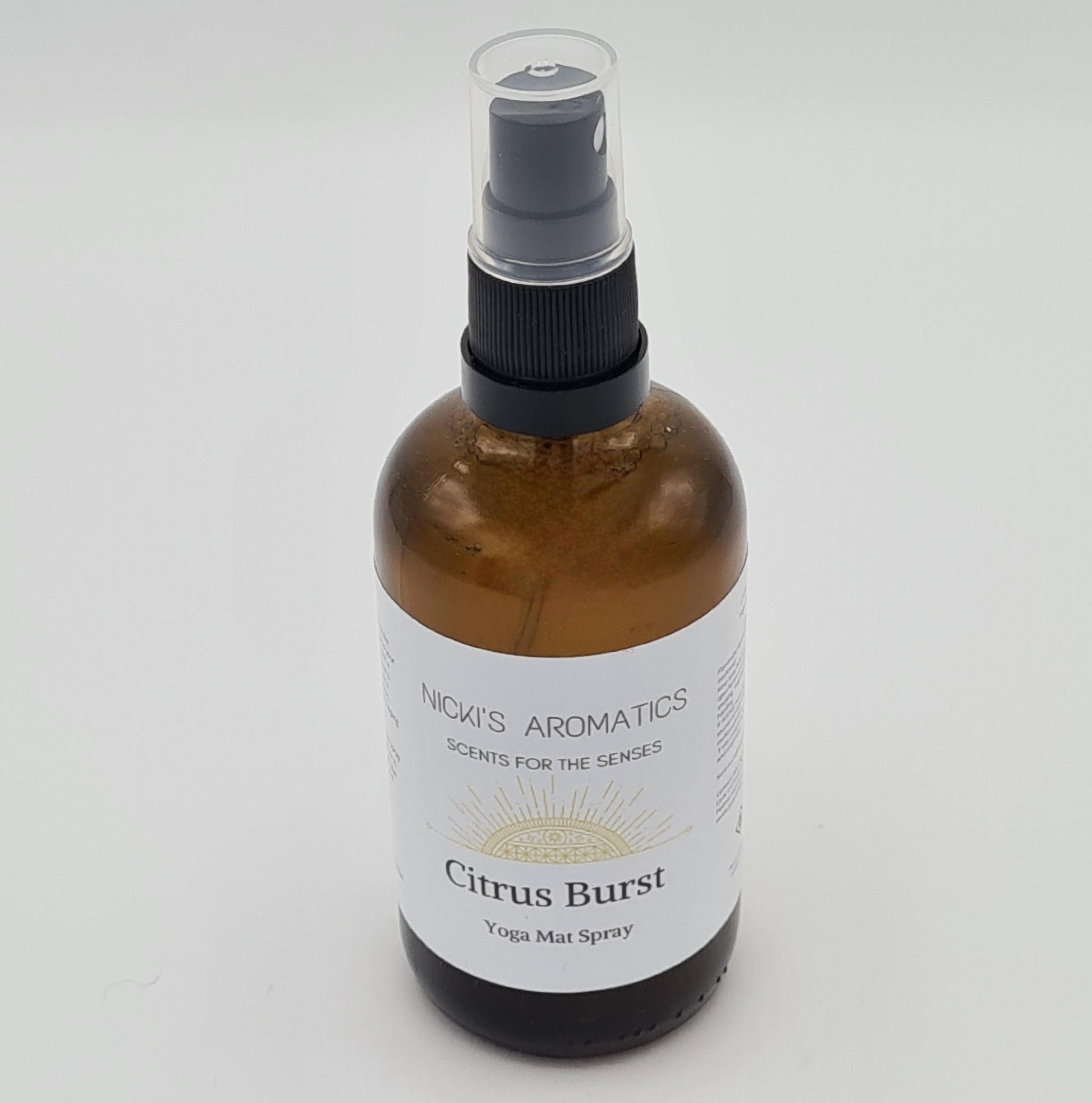 Amber glass spray bottle with clear plastic lid. At the top of the white label is the logo for "Nicki's Aromatics" with the strapline "Scents for the Senses" and at the bottom of the label is the text "Citrus Burst Yoga Mat Spray".