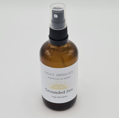 Amber glass spray bottle with clear plastic lid. At the top of the white label is the logo for "Nicki's Aromatics" with the strapline "Scents for the Senses" and at the bottom of the label is the text "Grounded Zen Yoga Mat Spray".