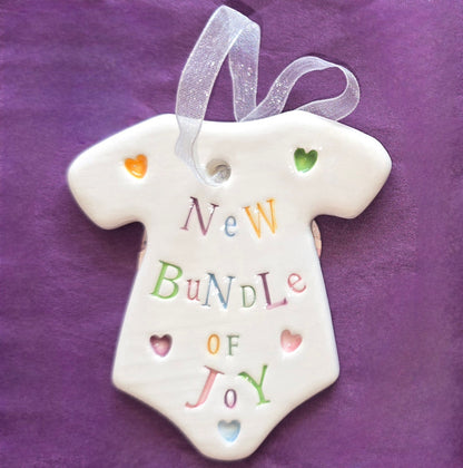 White ceramic baby grow shaped keepsake with the words "New Bundle of Joy" and some small heart illustrations dotted around. There is a white ribbon at the top of the heart for hanging.