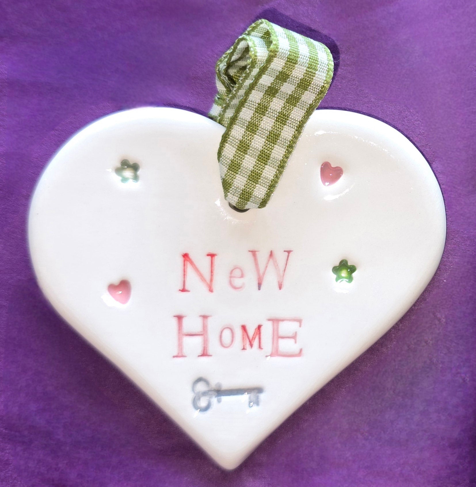 White ceramic heart with the words "New Home" and some small hearts, flowers and a key illustration dotted around. There is a green checked ribbon at the top of the heart for hanging.