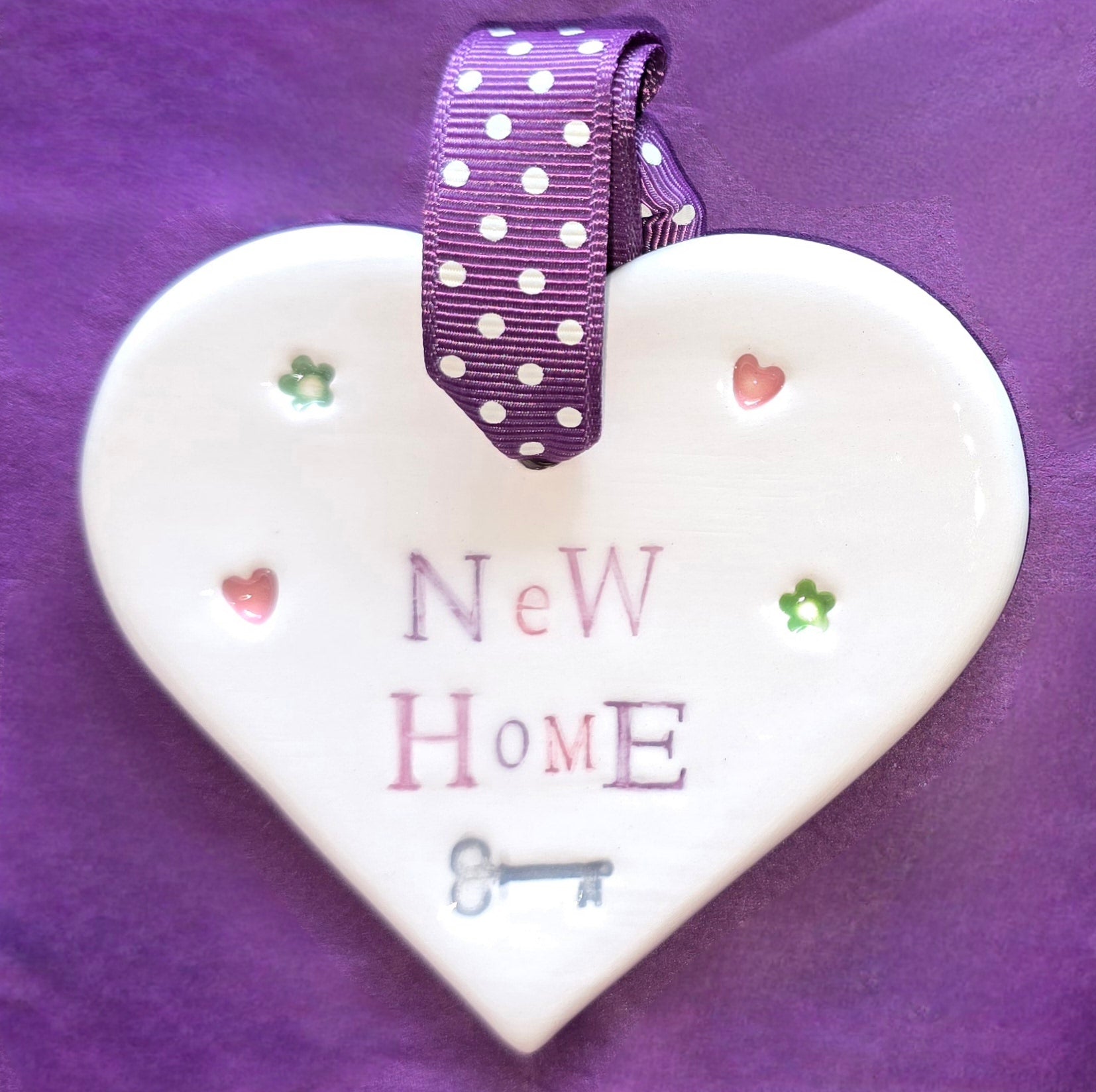 White ceramic heart with the words "New Home" and some small hearts, flowers and a key illustration dotted around. There is a purple polka dot ribbon at the top of the heart for hanging.