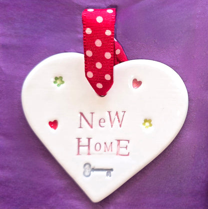 White ceramic heart with the words "New Home" and some small hearts, flowers and a key illustration dotted around. There is a red polka dot ribbon at the top of the heart for hanging.