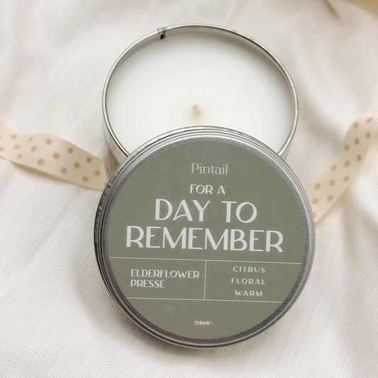 An open candle tin showing the inside of the candle with the lid overlaid. The label on the lid says "For A Day to Remember" with the text "Elderflower Presse" and "Citrus, Floral, Warm"