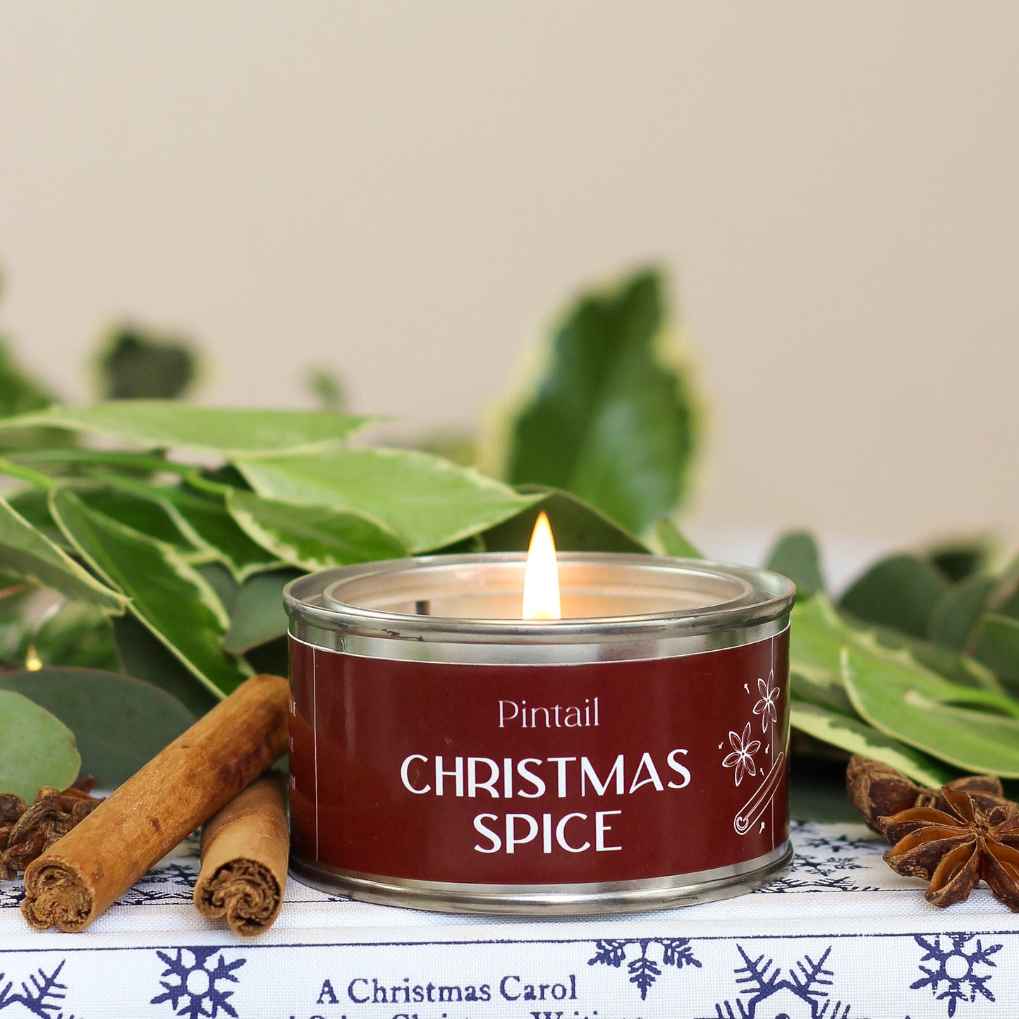 Paint Pot candle from Pintail with a brown label saying "Christmas Spice". The wick on the candle is burning and behind the candle there is some holly. To the left of the candle are two cinnamon sticks and to the right there are some cloves.