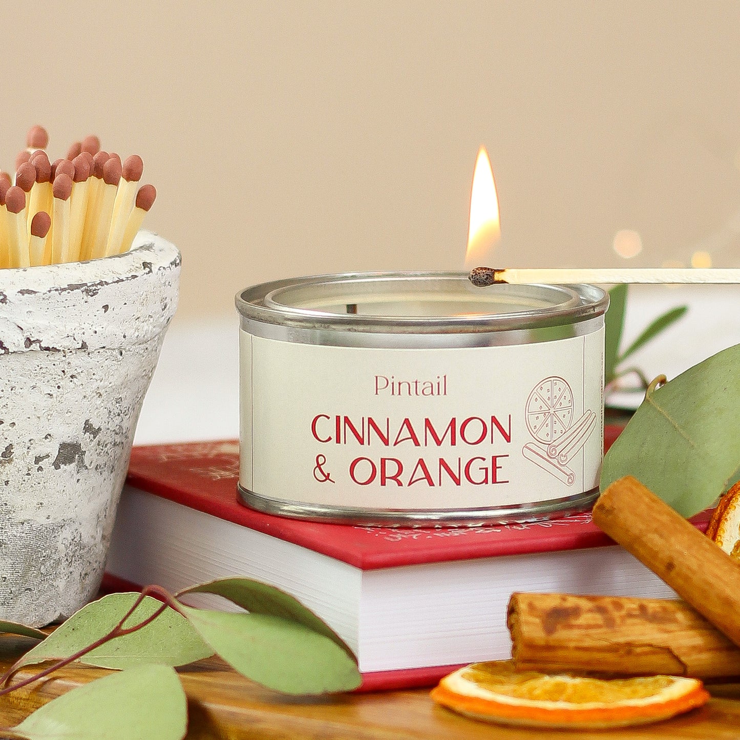 In the middle of the image there is a paint point tin candle being lit by a match. The cream coloured label on the candle says "Pintail" and "Cinnamon & Orange". The candle is sitting on top of a red book and to the left of this there is a white ceramic pot containing matches. To the right of the image, at the front, there are two cinnamon sticks and a slice of orange.