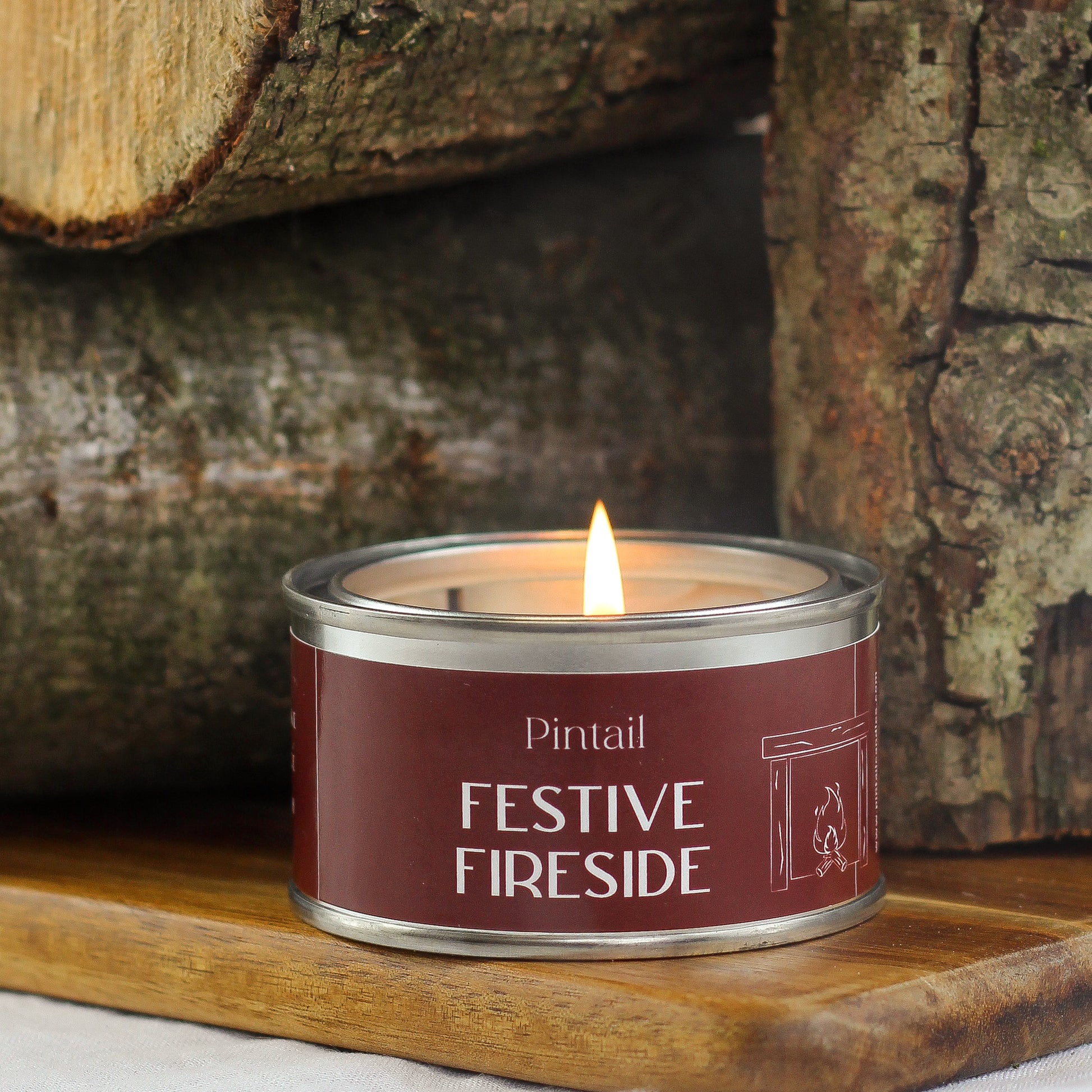 Pintal Festive Fireside paint pot candle with a brown label sitting on a  wooden shelf. The wick on the candle is lit and behind the candle there are three logs, two piled one on top of the other with the third log standing upright to the right of the others.