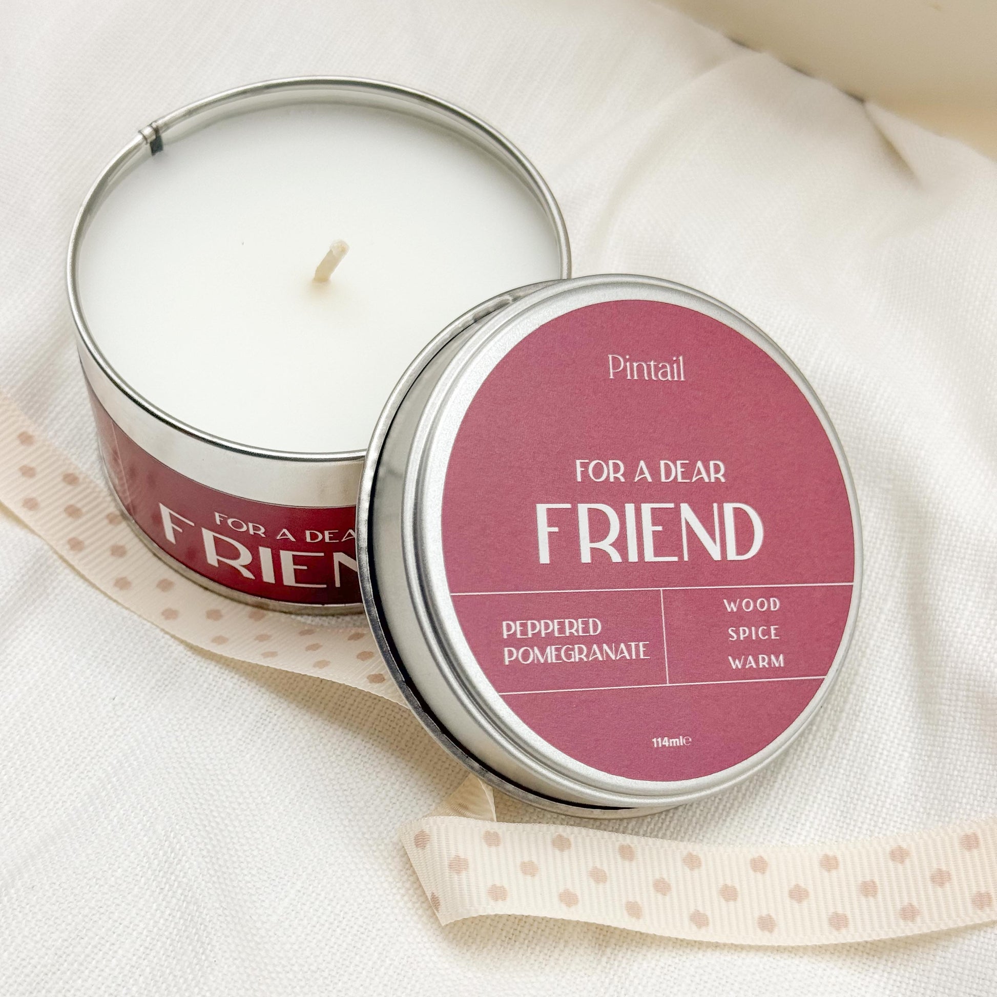 View from above of open candle tin from Pintail Candles showing the white candle on the left hand side of image and the tin lid leaning on the tin on the right hand side of the image. The wording on the lid says "For a Dear Friend", "Peppered Pomegranate", "Wood, Spice, Warm".