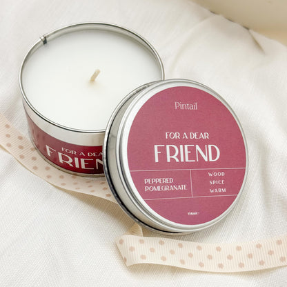 View from above of open candle tin from Pintail Candles showing the white candle on the left hand side of image and the tin lid leaning on the tin on the right hand side of the image. The wording on the lid says "For a Dear Friend", "Peppered Pomegranate", "Wood, Spice, Warm".