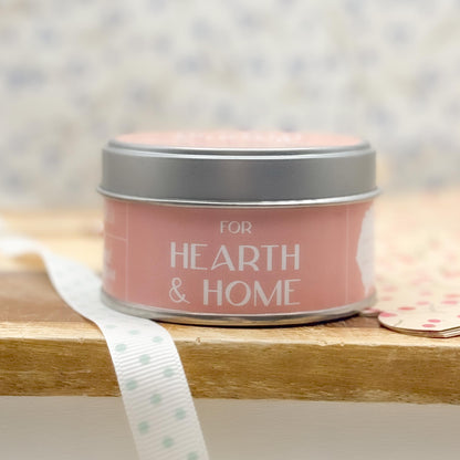 Pink candle tin with silver lid sitting on a wooden shelf or table. Label says "For Hearth & Home". 