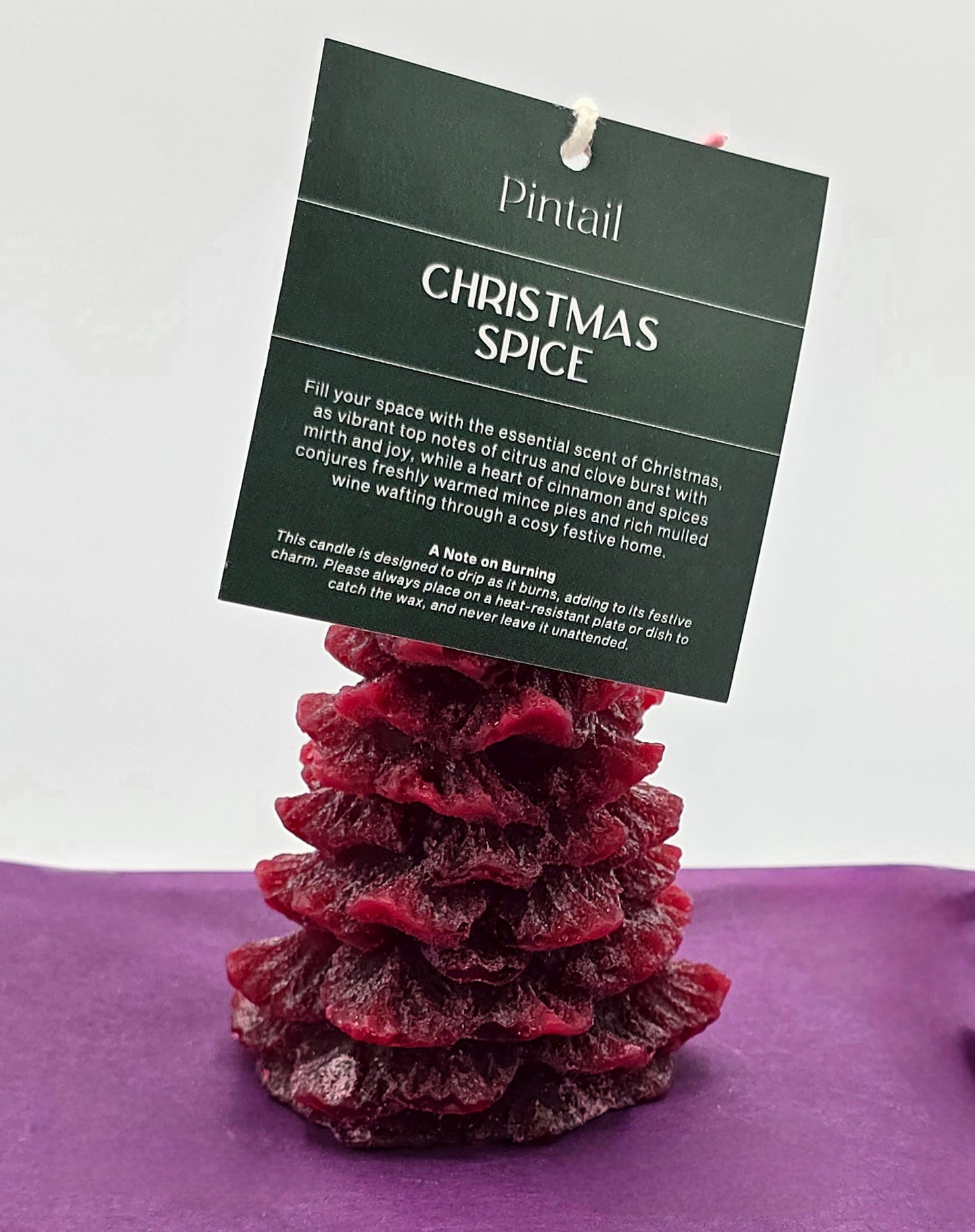 Red candle shaped like a Christmas tree with a 'Pintail' Christmas Spice label on a purple surface.