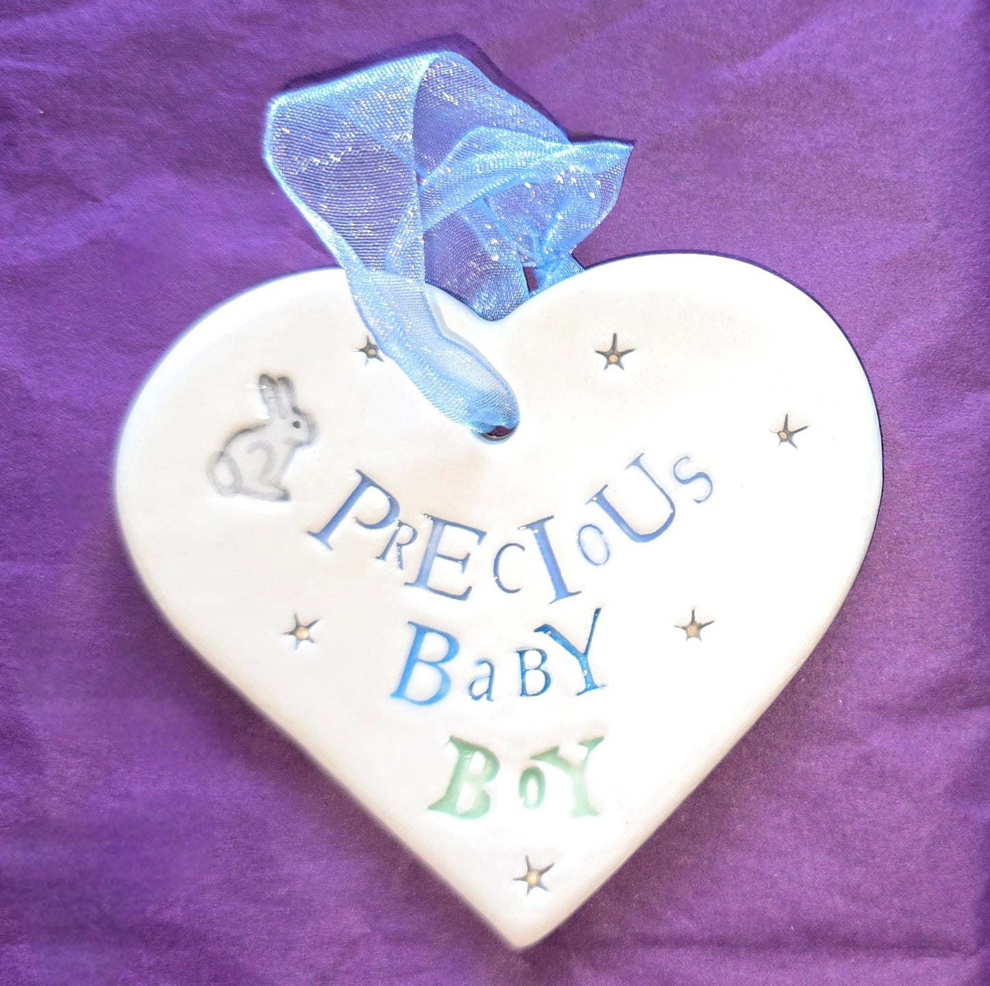 White ceramic heart with the words "Precious Baby Boy" and some small star shaped illustrations and a small grey bunny illustration dotted around. There is a pale blue ribbon at the top of the heart for hanging.