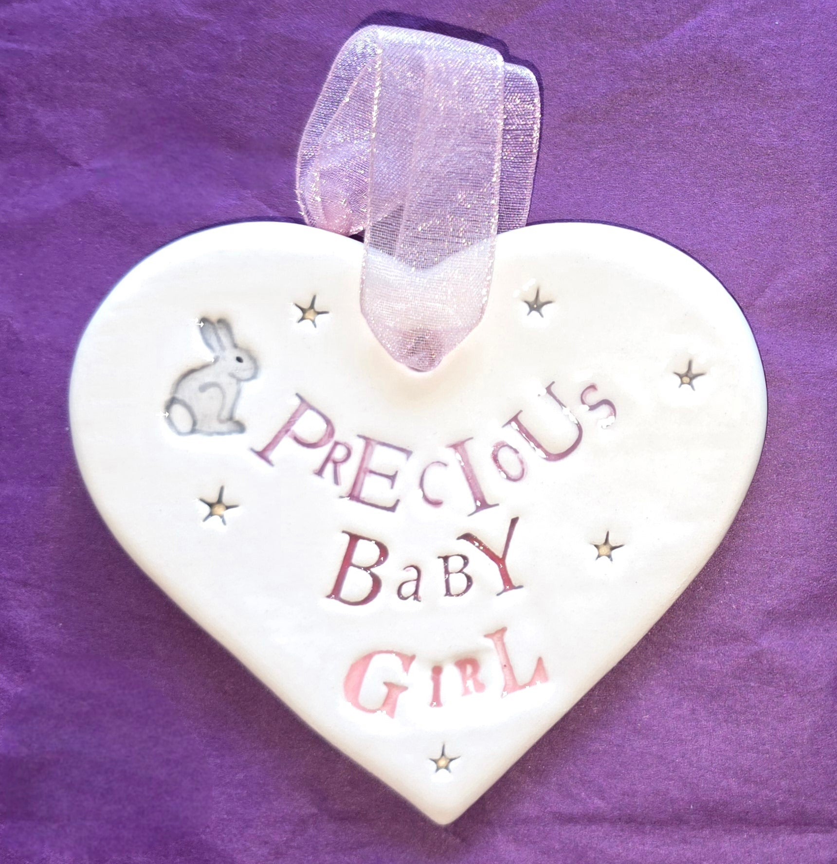 White ceramic heart with the words "Precious Baby Girl" and some small star shaped illustrations and a small grey bunny illustration dotted around. There is a pale pink ribbon at the top of the heart for hanging.