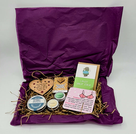 Letterbox friendly cardboard box lined with purple tissue paper and brown shredded tissue. The box contains a heart shaped wooden plaque, a Seedball tin, small tins of lip balm and nail & cuticle balm, a coaster, a little pocket hug and a card with the slogan "I Miss Your Face". 