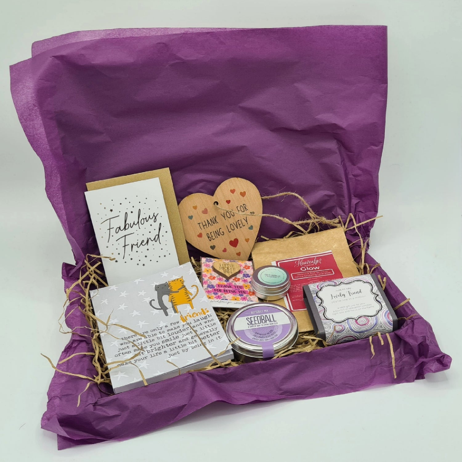 Lovely Friend Letterbox Gift - a letterbox friendly cardboard box lined with purple tissue paper and brown shredded tissue. It contains an A6 notebook, a "Fabulous Friend" mini card, a heart shaped wooden plaque, a Little Pocket Hug, a Seedball tin, a tin of Lip Balm, a Glow facial mask and Lovely Friend mini candles.