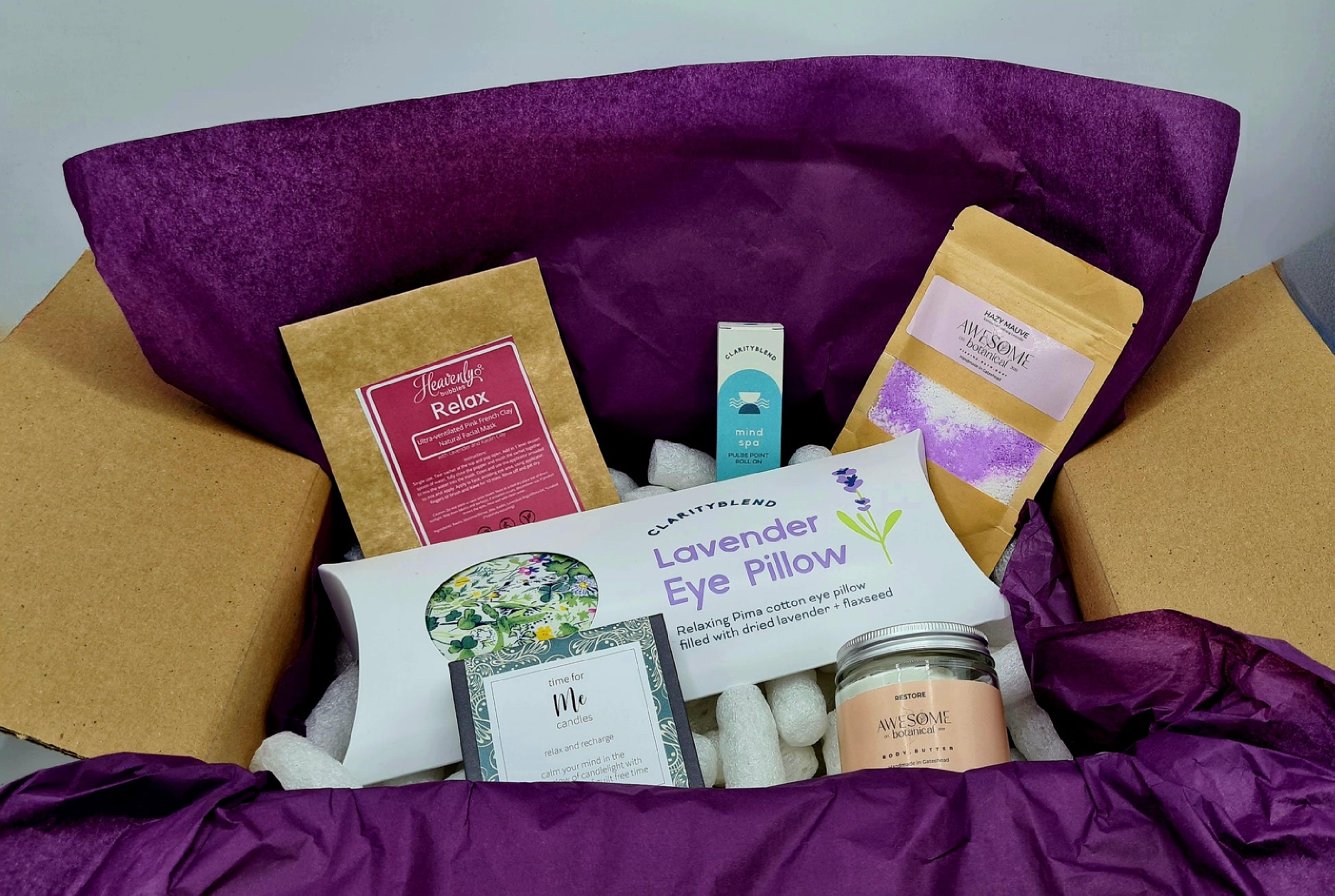 Restore & Relax Giftbox - brown cardboard box lined with purple tissue paper and white biodegradable packing peanuts. The box contains Relax facial mask from Heavenly Bubbles; Lavender Eye Pillow and Mind Spa Pulse Point Roll On from Clarity Blend; Hazy Mauve Bath Dust and Restore Body Butter from Awesome Botanical and box of 12 mini candles with the message "Time for Me". 