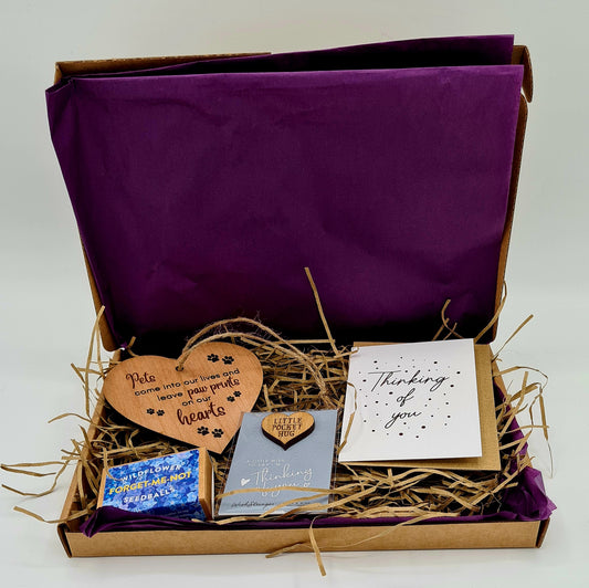 Letterbox friendly brown cardboard box lined with purple tissue paper and brown shredded tissue. The box contains a heart shaped wooden plaque with the slogan "Pets come into our lives and leave paw prints on our hearts"; match box sized box from Seedball containing Forget-Me-Not seeds; Little Pocket Hug with the message "Thinking of You" and a mini card with the message "Thinking of You". 