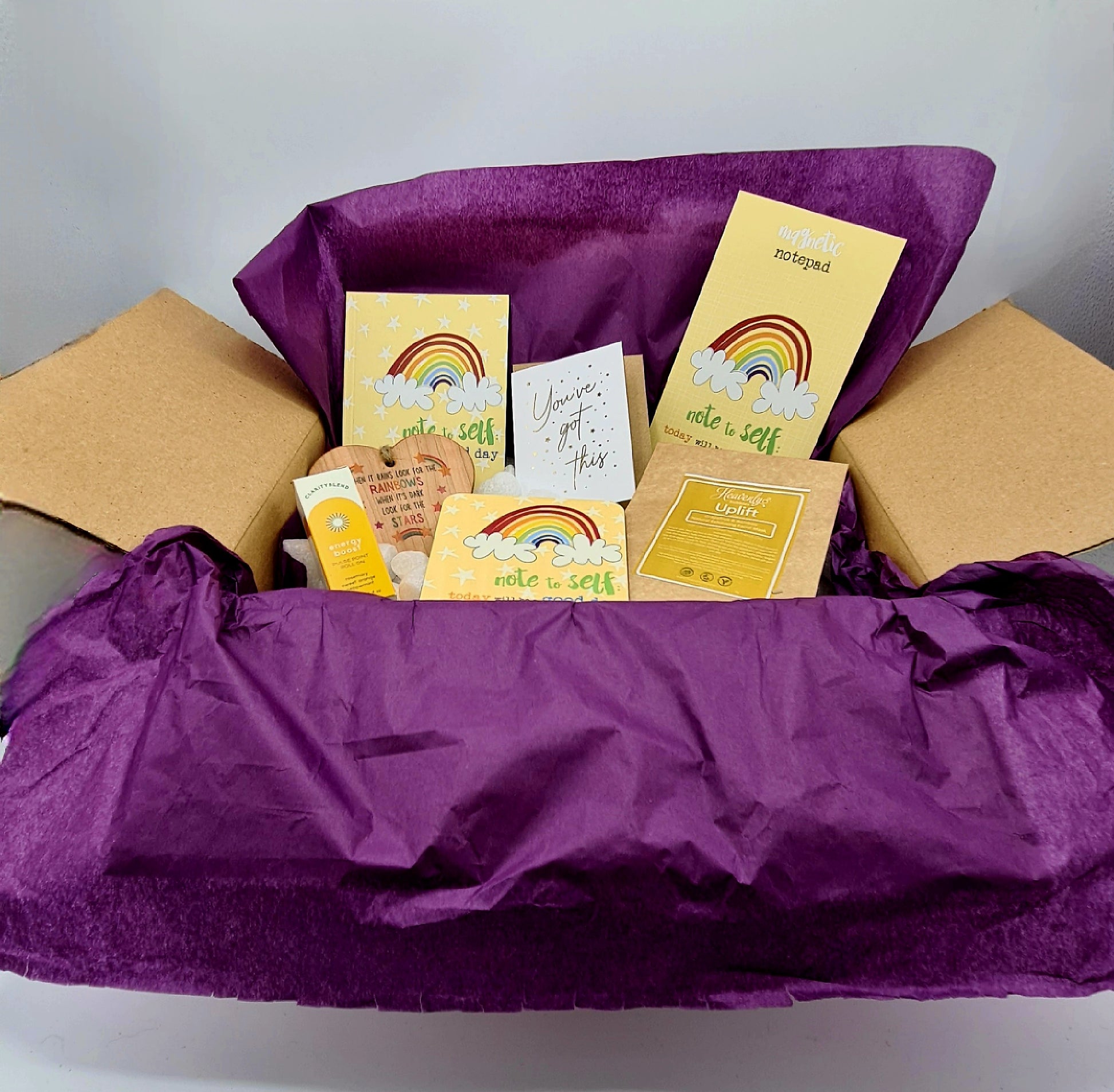 Open brown cardboard gift box lined with purple tissue paper and white biodegradable packing peanuts. It contains various yellow coloured items including A6 Notebook, Magnetic Notepad and Coaster from Dandelion Stationery with the slogan "Note to Self: Today will be a good day"; Energy Boost Pulse Point Roll On from Clarity Blend; Uplift Facial Mask from Heavenly Bubbles; heart shaped wooden plaque and mini card with the message "You've Got This". 