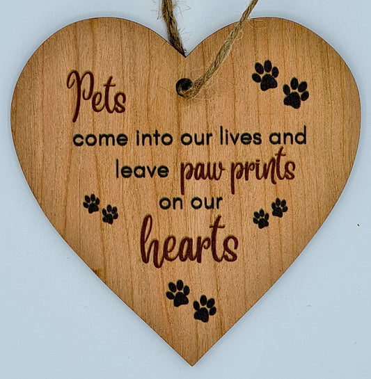 Heart shaped wooden plaque with the slogan "Pets come into our lives and leave paw prints on our hearts". There are several black paw prints around the writing. At the top of the plaque there is a jute string for hanging.