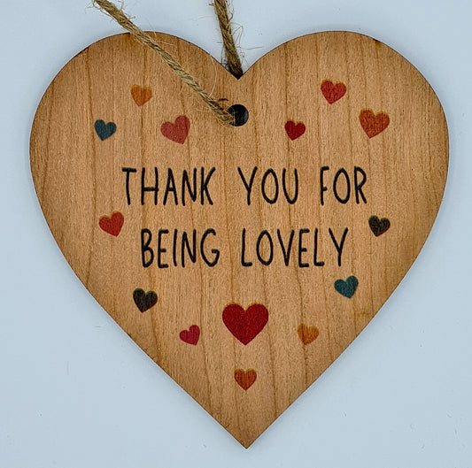 Heart shaped wooden plaque with the slogan "Thank you for being lovely". Around the writing, there are lots of little hearts in red, orange, blue and black. There is a jute string for hanging at the top of the plaque.