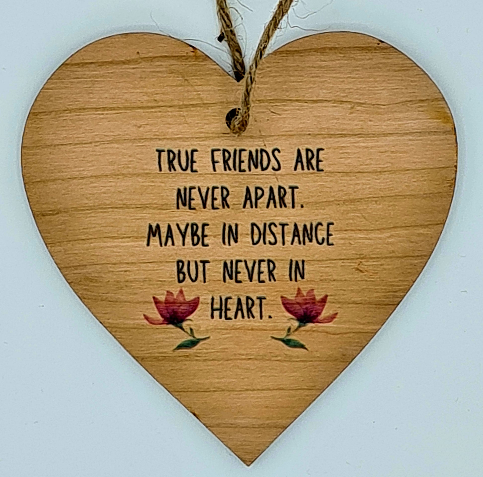 Heart shaped wooden plaque with the slogan "True friends are never apart. Maybe in distance but never in heart". There are two flowers underneath the writing. At the top of the plaque, there is a jute string for hanging.