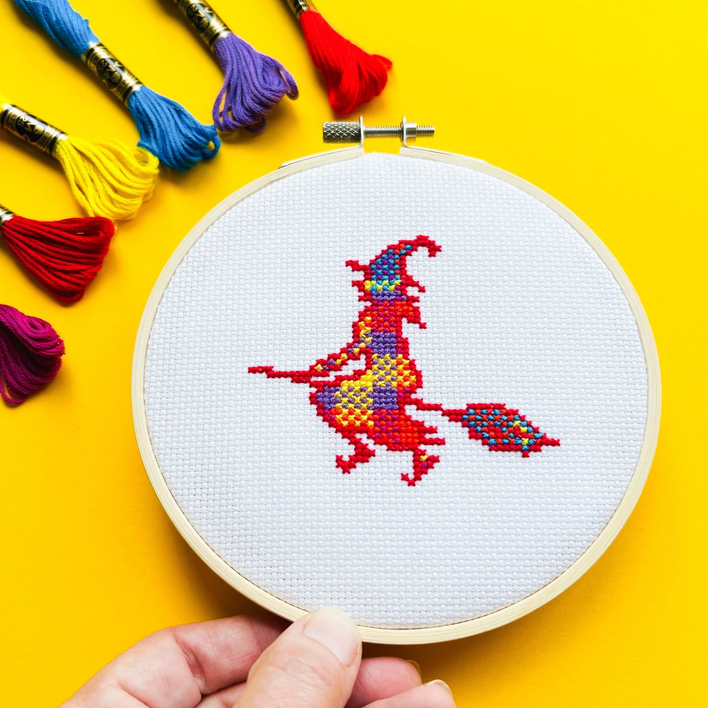 Bright yellow background with six brightly coloured threads in the top left corner and a round embroidery hoop in the middle of the picture. Inside the embroidery hoop there is a cross stitched Agnes the Witch.