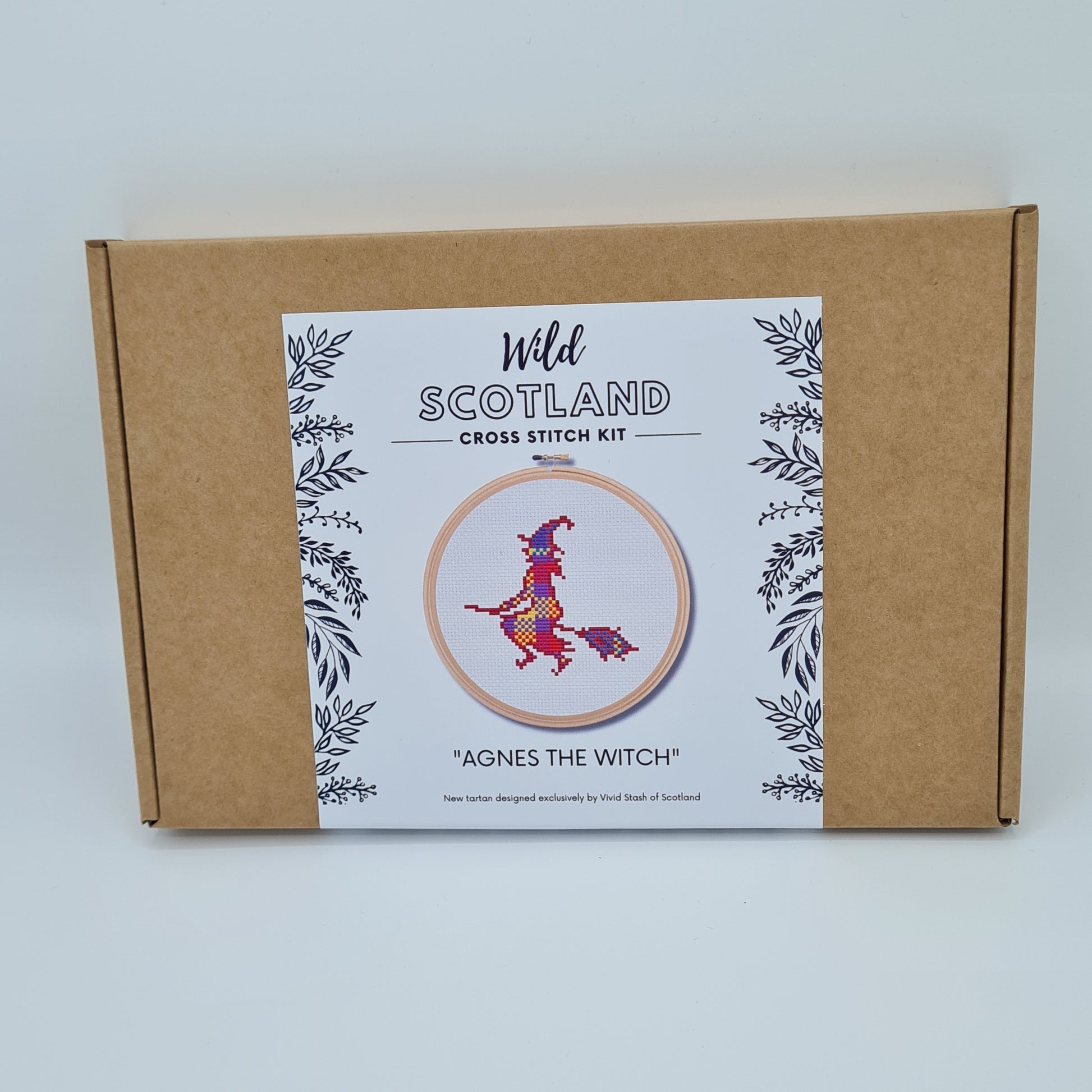 Brown cardboard box with white label saying Wild Scotland Cross Stitch Kit. Below the name, there is a picture of an embroidery hoop containing a cross stitched witch. Below the image, there is the title "Agnes the Witch" with text below saying "New tartan designed exclusively by Vivid Stash of Scotland".