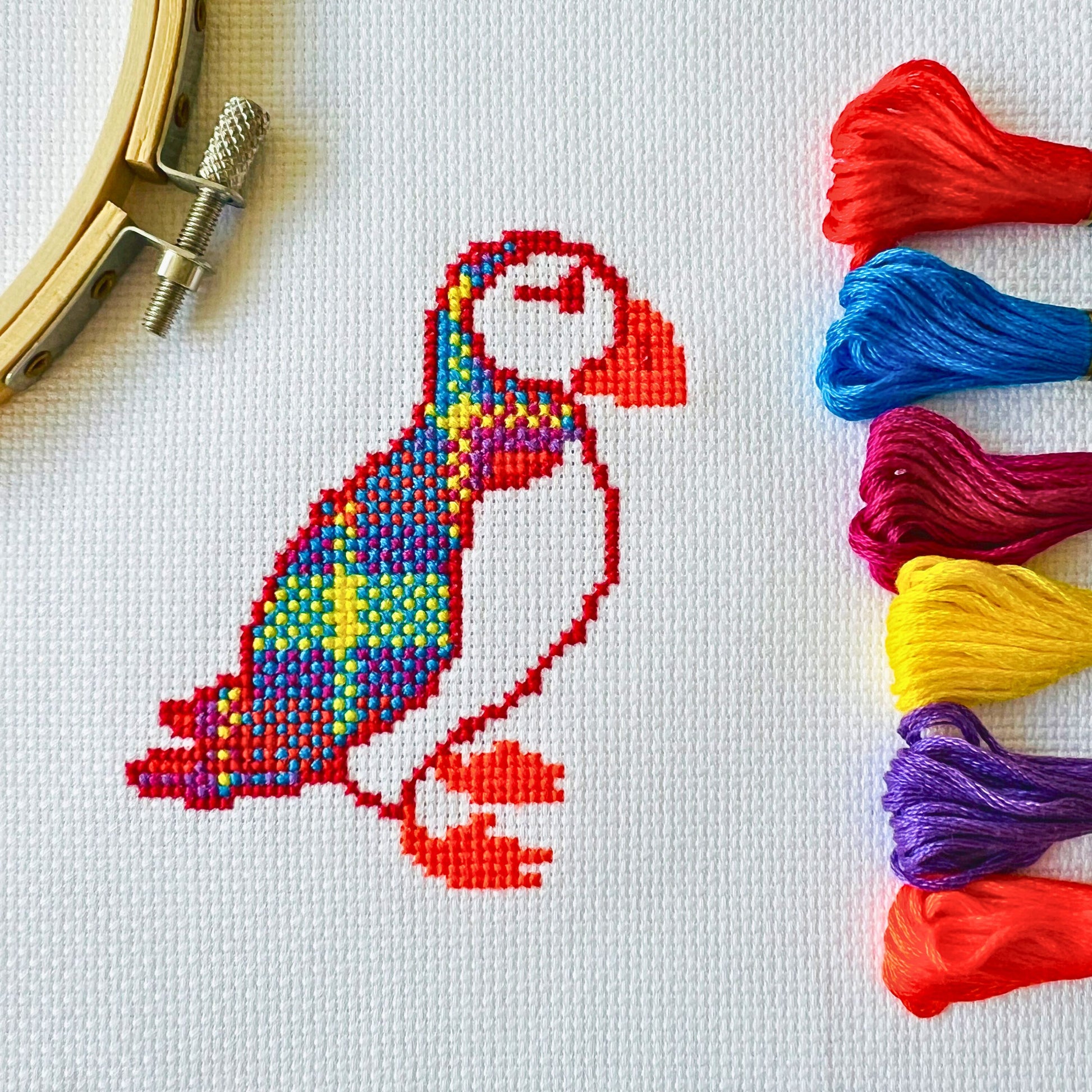 White coloured fabric containing a cross stitched puffin in the bright colours of the Vivid Stash tartan. To the right of the puffin there are six skeins of thread starting from red to blue to pink to yellow to purple to orange as you look from top to bottom.