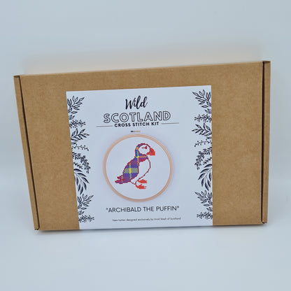 Kraft brown flat cardboard box with a white label saying Wild Scotland Cross Stitch Kit. Below the name, there is a picture of an embroidery hoop containing a cross stitched puffin. Below the image, there is the title "Archibald the Puffin" with text below saying "New tartan designed exclusively by Vivid Stash of Scotland".