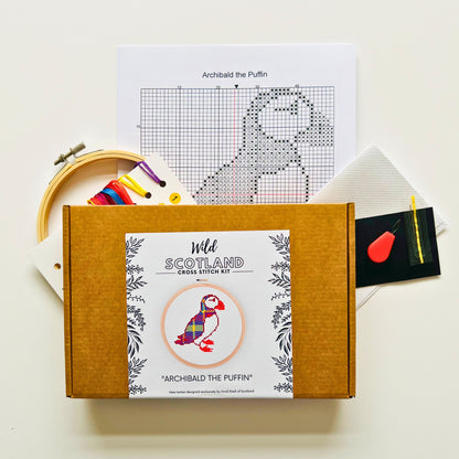 Image showing the contents of the Cross Stitch Kit with a chart entitled Archibald the Puffin at the top. To the left of this, there is an embroidery hoop, overlaid by a card of threads. To the right there is a folded piece of fabric, a needle and a needle threader. At the bottom of the image, overlaid is a kraft brown flat cardboard box with a label saying "Wild Scotland Cross Stitch Kit", an image of a cross stitched puffin inside an embroidery hoop with a title below stating "Archibald the Puffin".