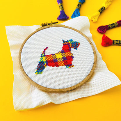 Bright yellow background with five skeins of brightly coloured threads in the top right corner and a round embroidery hoop in the middle of the picture. Inside the embroidery hoop there is a cross stitched tartan Blair the Scottie Dog.