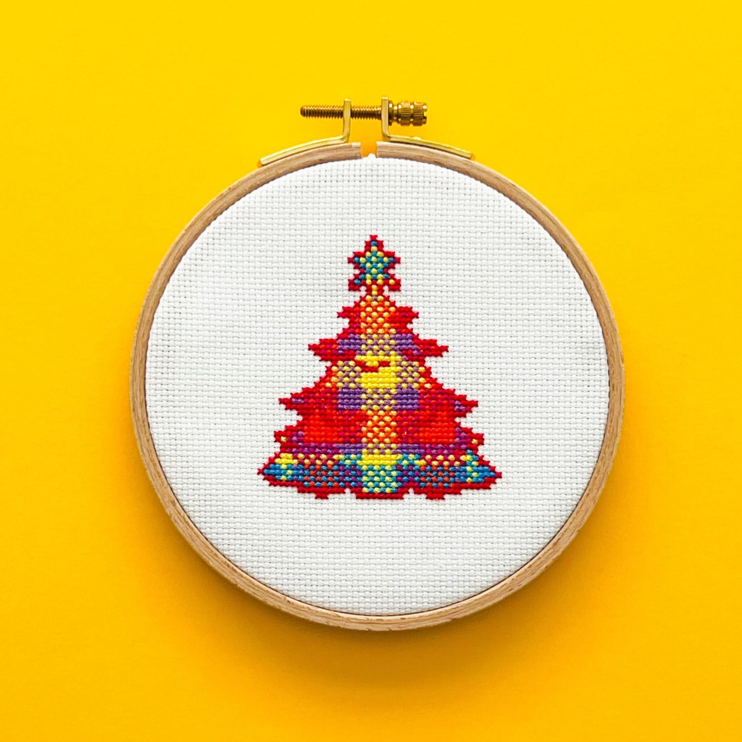 Bright yellow background with a round embroidery hoop in the middle of the picture. Inside the embroidery hoop there is a cross stitched tartan Christmas Tree.