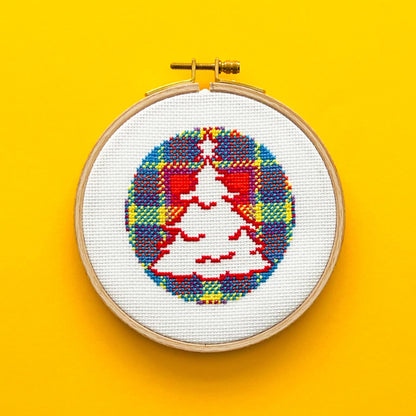 Bright yellow background with a round embroidery hoop in the middle of the picture. Inside the embroidery hoop there is a cross stitched Christmas Tree Encircled in the Vivid Stash Tartan.