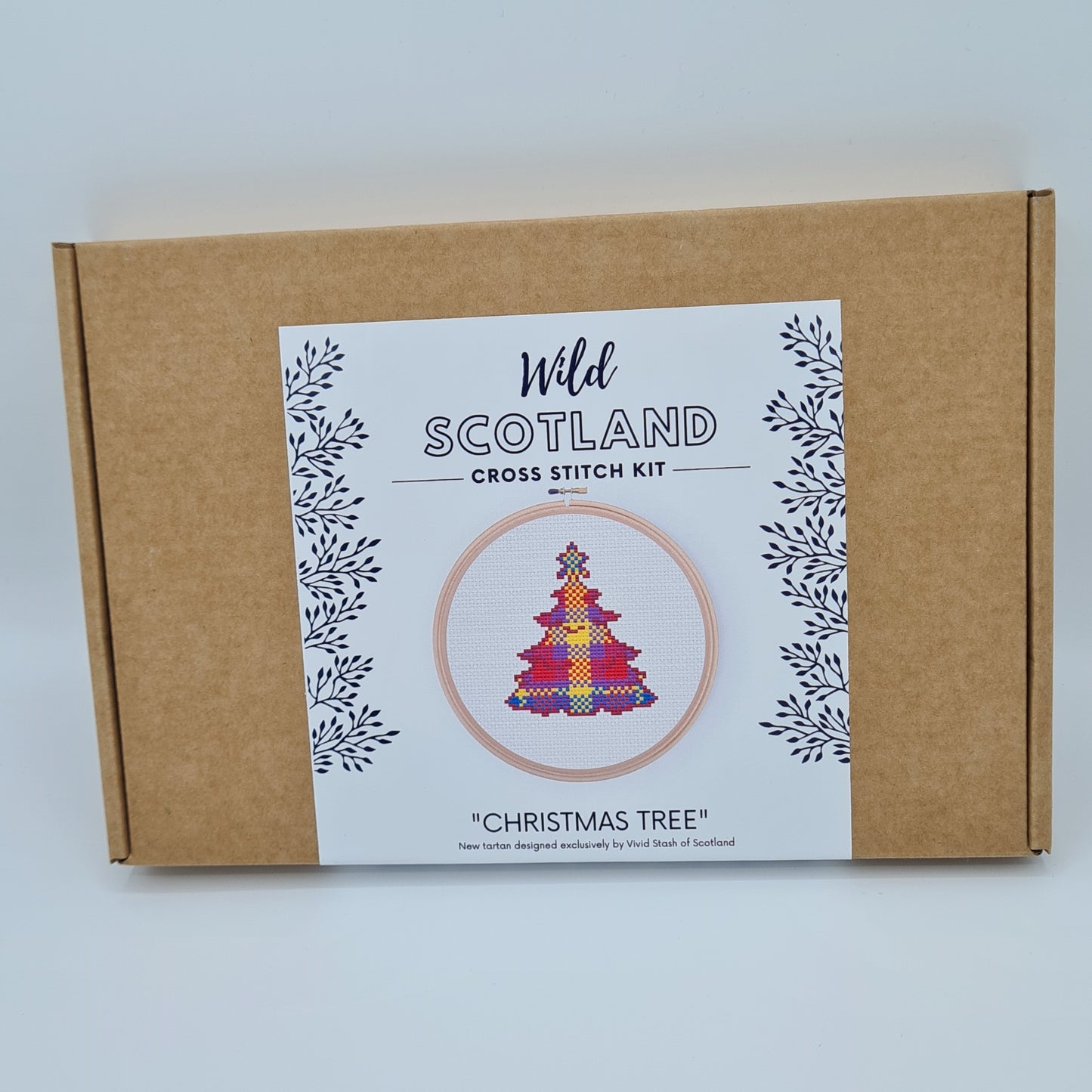 Kraft brown flat cardboard box with a white label saying Wild Scotland Cross Stitch Kit. Below the name, there is a picture of an embroidery hoop containing a cross stitched Christmas tree in tartan. Below the image, there is the title "Christmas Tree" with text below saying "New tartan designed exclusively by Vivid Stash of Scotland".