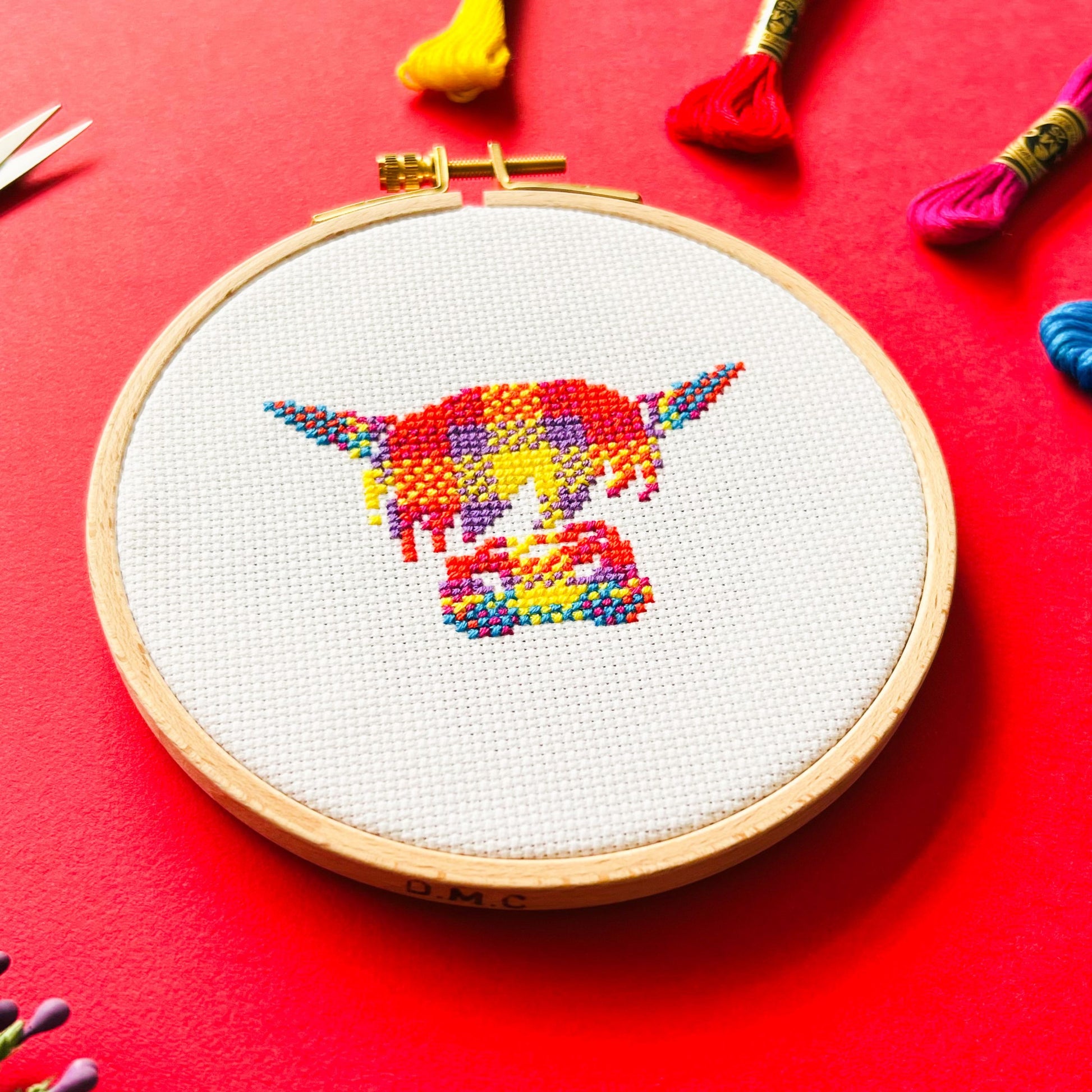 Bright red background with four brightly coloured skeins of thread in the top rightt corner and a round embroidery hoop in the middle of the picture. Inside the embroidery hoop there is a cross stitched Highland Cow in brightly coloured tartan.