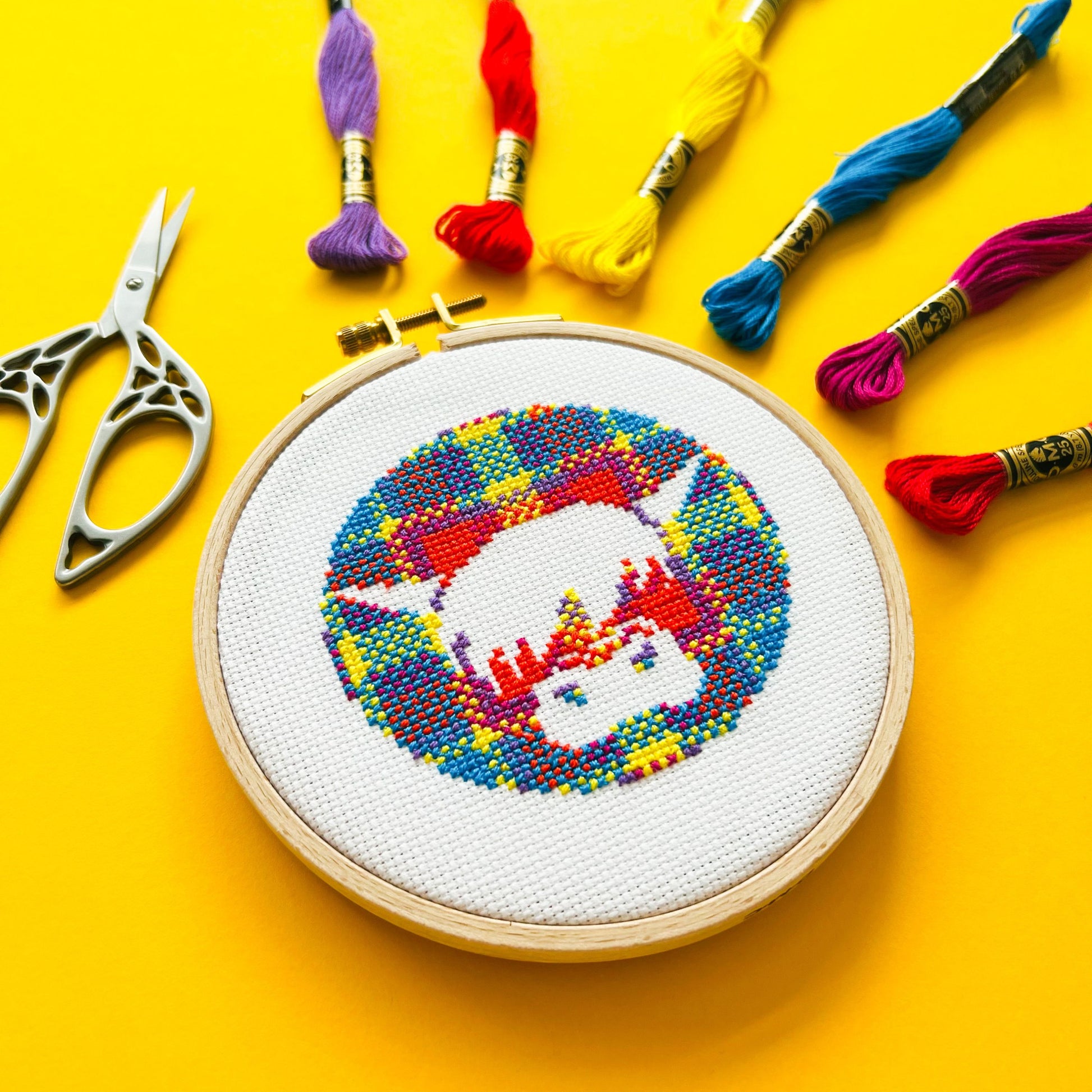 Bright yellow background with six brightly coloured skeins of thread in the top right corner, scissors on the left and a round embroidery hoop in the middle of the picture. Inside the embroidery hoop there is a cross stitched Harris the Highland Cow, Encircled in tartan.