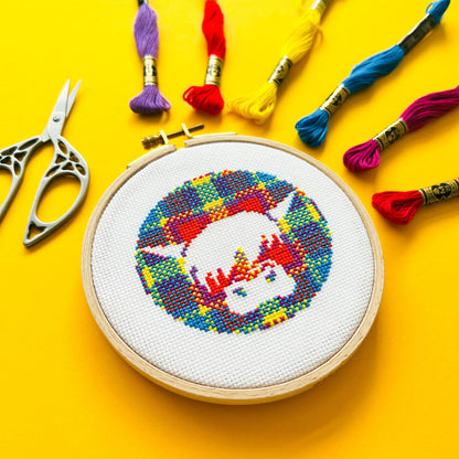 Bright yellow background with six brightly coloured skeins of thread in the top right corner, scissors on the left and a round embroidery hoop in the middle of the picture. Inside the embroidery hoop there is a cross stitched Harris the Highland Cow, Encircled in tartan.