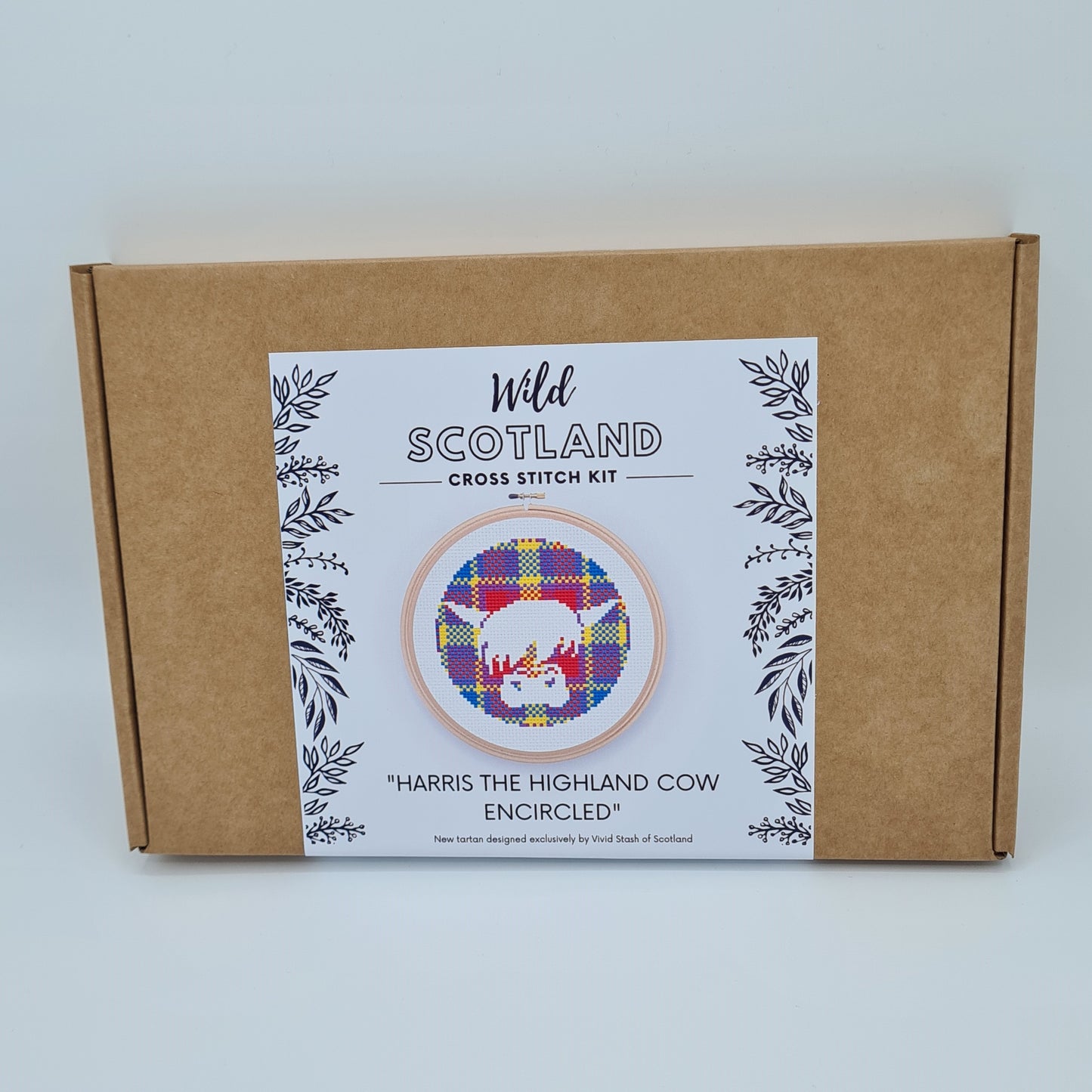 Kraft brown flat cardboard box with a white label saying Wild Scotland Cross Stitch Kit. Below the name, there is a picture of an embroidery hoop containing a cross stitched face of a Highland Cow, encircled in the Vivid Stash tartan. Below the image, there is the title "Harris the Highland Cow Encircled" with text below saying "New tartan designed exclusively by Vivid Stash of Scotland".