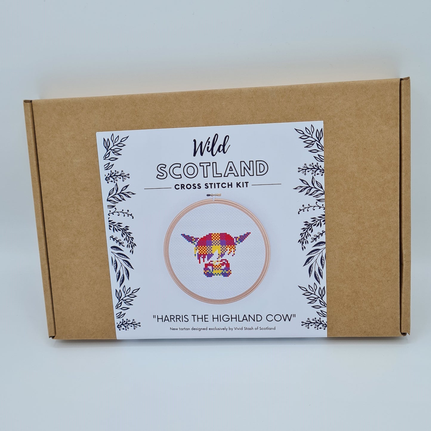 Kraft brown flat cardboard box with a white label saying Wild Scotland Cross Stitch Kit. Below the name, there is a picture of an embroidery hoop containing a cross stitched face of a Highland Cow. Below the image, there is the title "Harris the Highland Cow" with text below saying "New tartan designed exclusively by Vivid Stash of Scotland".