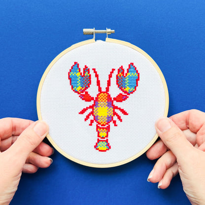 Bright blue background with two hands at the bottom of the image holding an embroidery hoop. The embroidery hoop contains a cross stitched image of Larry the Lobster.