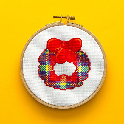 Bright yellow background with a round embroidery hoop in the middle of the picture. Inside the embroidery hoop there is a cross stitched Tartan Wreath with a red bow at the top of it.